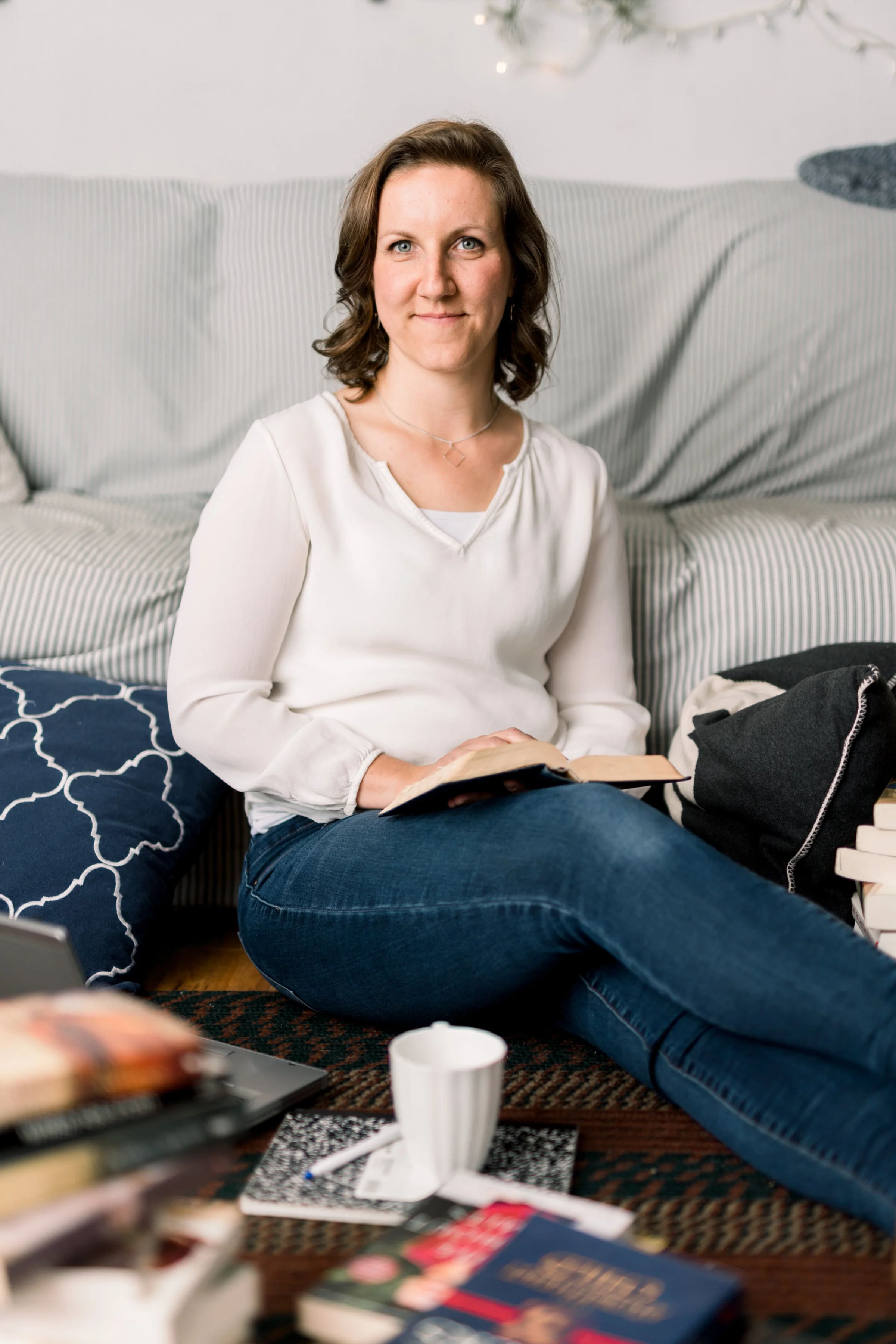 An author sitting on the floor by a couch with an open book and stacks of novels around her.